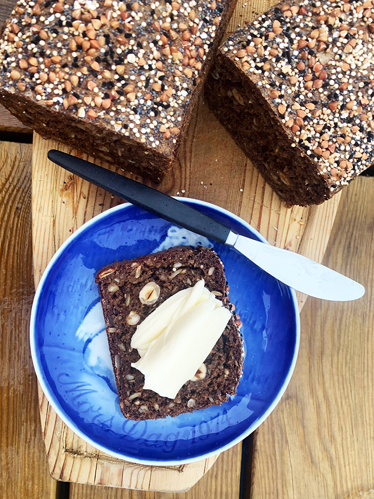 Rugbrød med hasselnødder, billedet er taget oppe fra og man ser rugbrødets top spækket med kerner og ved siden af en blå tallerken med en skive nybagt rugbrød med tandsmør